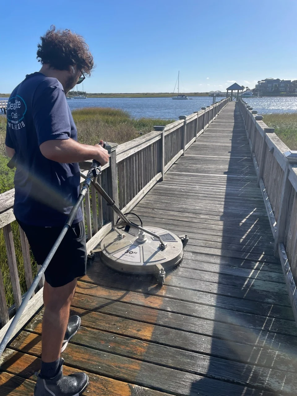 06e2f14abd42de48bc4191705531fc5bd6e0fef36b6efbae96065a81988049e8 Person pressure washing a wooden boardwalk by the water in Wilmington, NC, clearing dirt and restoring the surface.