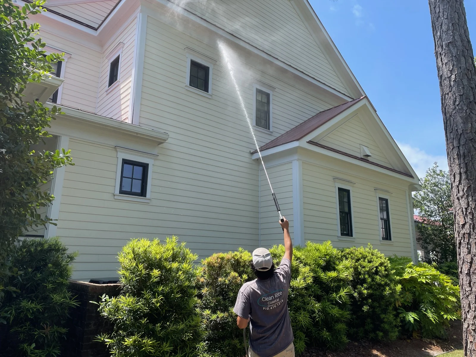 Person pressure washing the exterior of a large, light yellow house surrounded by greenery, representing pressure washing Wilmington NC.