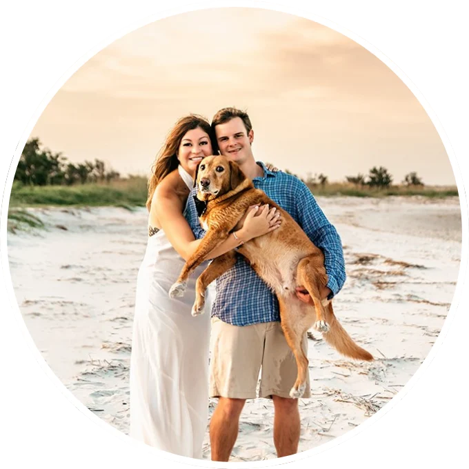Couple on sandy beach smiling, holding a large brown dog, under an overcast sky, evoking a serene atmosphere akin to pressure washing Wilmington NC.