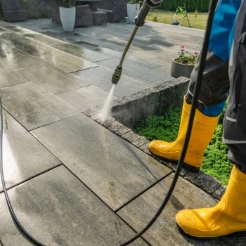 Porch & Concrete Cleaning Person in yellow boots uses a pressure washer on a patio. Clean patio stones gleam under the sun. Pressure washing Wilmington NC.