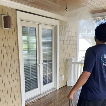 Person pressure washing a wooden porch, focusing on glass doors, in Wilmington, NC. Light mist visible near the ceiling.