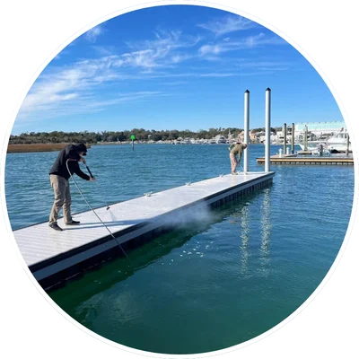Two people pressure washing a dock in Wilmington, NC, surrounded by water and boats under a clear blue sky.